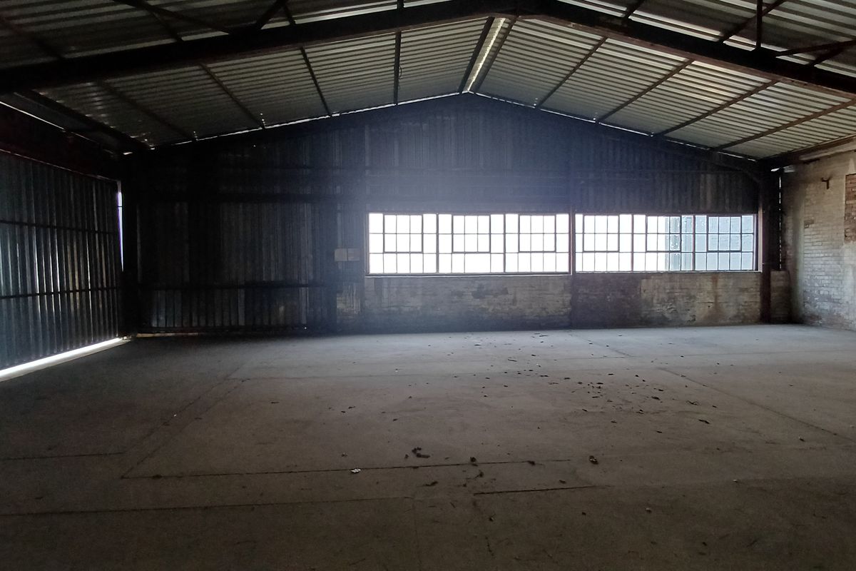 Spacious industrial warehouse interior featuring exposed steel roof trusses, concrete flooring, and large steel-framed windows providing natural light.