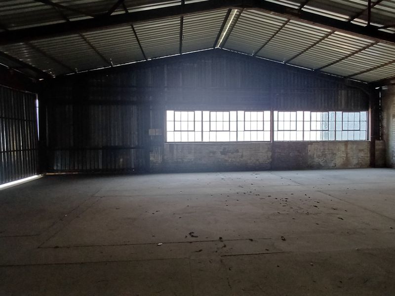 Spacious industrial warehouse interior featuring exposed steel roof trusses, concrete flooring, and large steel-framed windows providing natural light.