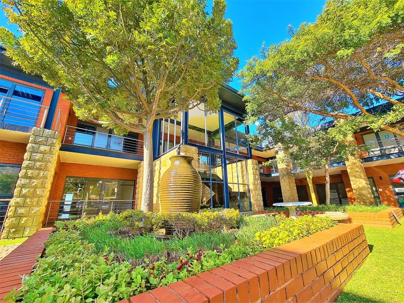 A well-maintained office complex exterior featuring a central courtyard with a water feature, landscaped gardens, and a modern brick and glass facade.