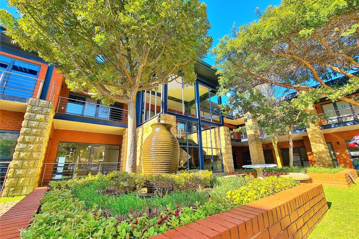A well-maintained office complex exterior featuring a central courtyard with a water feature, landscaped gardens, and a modern brick and glass facade.