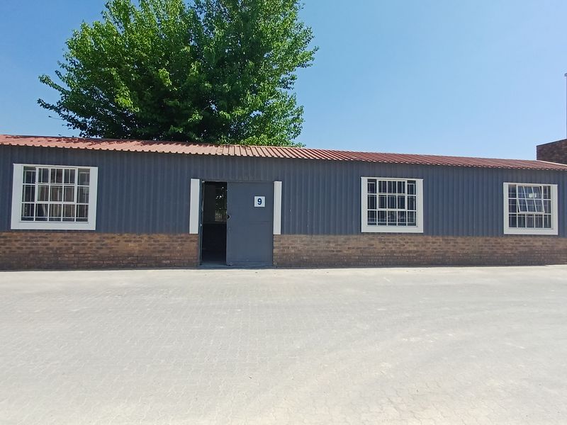 Exterior view of a single-story office building featuring corrugated metal cladding, a brick base, and secure window bars, set on a paved lot.