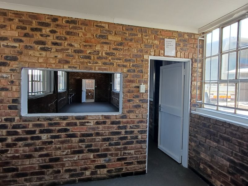 An empty office space featuring exposed brick walls, large steel-framed windows with security bars, and an internal service window connecting to an adjacent room.