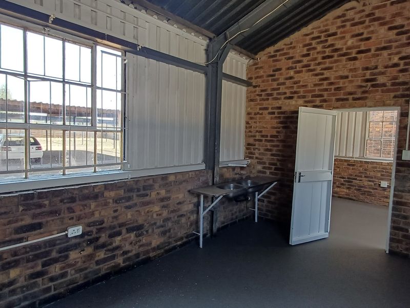 An open-plan office space featuring exposed brick walls, steel-framed windows, and a functional stainless steel sink area with industrial-style exposed roof trusses.