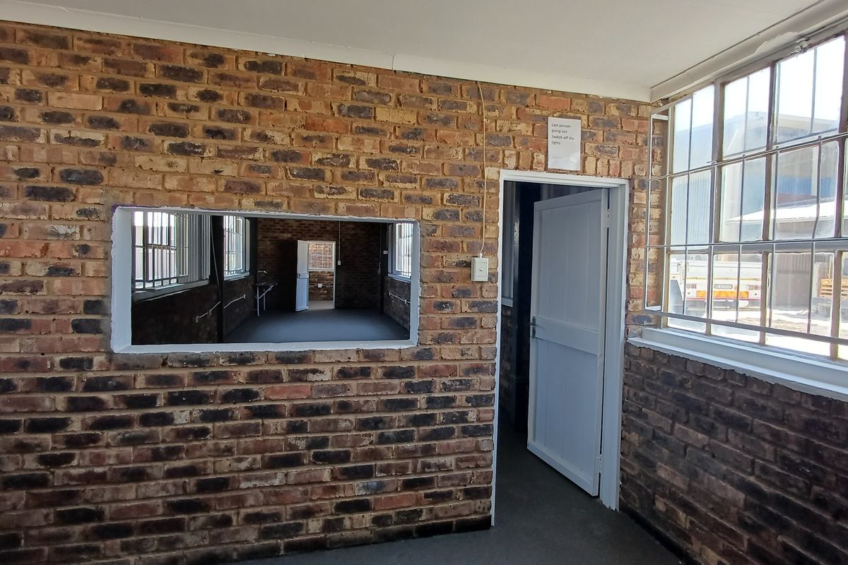 An empty office space featuring exposed brick walls, large steel-framed windows with security bars, and an internal service window connecting to an adjacent room.