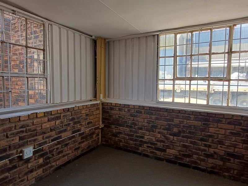 An empty office space featuring exposed brick walls, corrugated metal paneling, and large steel-framed windows with security bars.