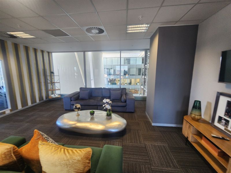 A modern office lounge area featuring floor-to-ceiling windows, suspended ceiling tiles, and carpeted flooring in a professional corporate environment.