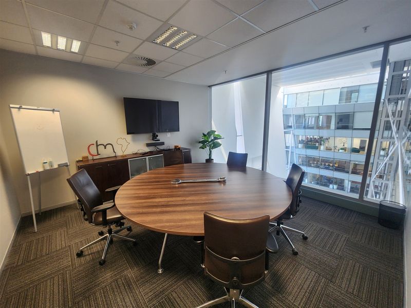 A professional office meeting room featuring a large round table, floor-to-ceiling windows with an atrium view, and modern carpet tile flooring.