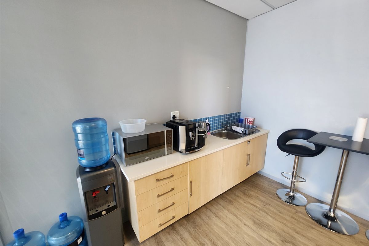 A compact office kitchenette featuring light wood cabinetry, a stainless steel sink, tiled backsplash, and durable laminate flooring.