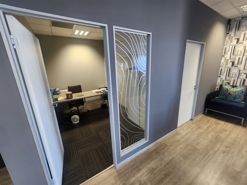 A modern office space featuring a private office room with carpet tiles, glass partition detailing, and a bright, professional hallway with wood-look laminate flooring.