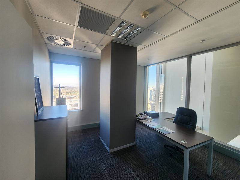 A professional office space featuring carpet tiles, a central structural column, suspended ceilings with integrated lighting, and large windows offering expansive city views.