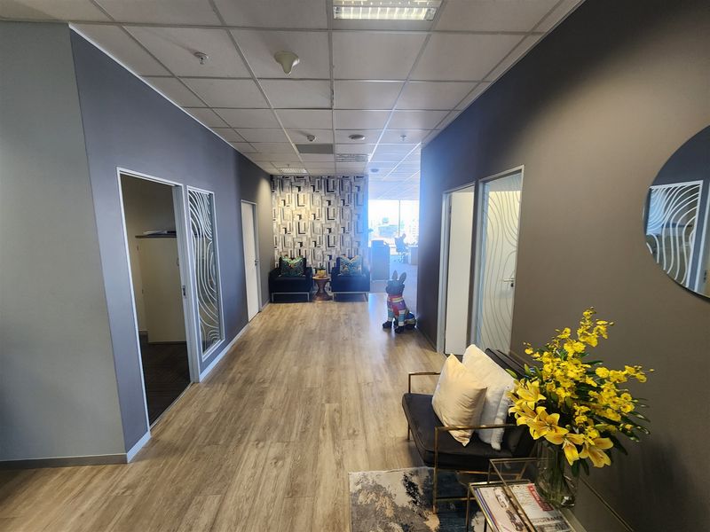 A modern office hallway featuring laminate flooring, suspended ceilings with recessed lighting, and glass-paneled doors leading to private office spaces.