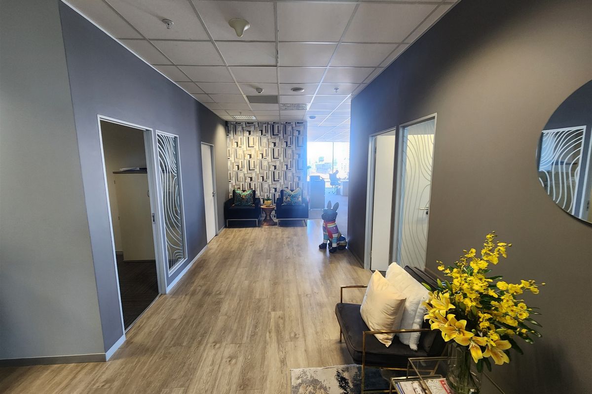 A modern office hallway featuring laminate flooring, suspended ceilings with recessed lighting, and glass-paneled doors leading to private office spaces.