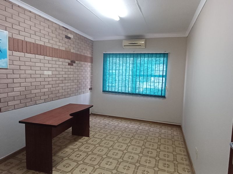 A functional office space featuring a brick accent wall, tiled flooring, an integrated air conditioning unit, and vertical window blinds.