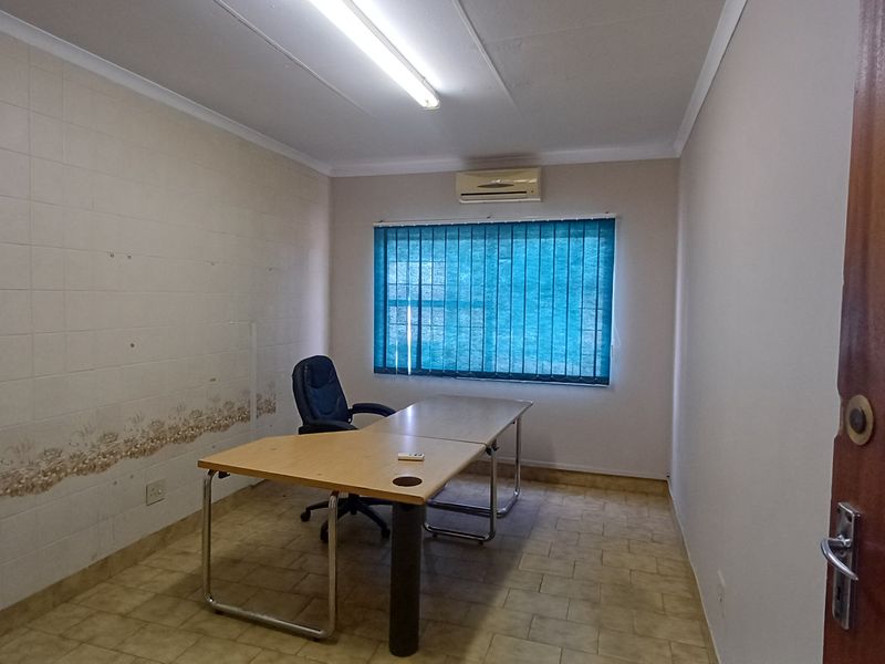 A functional office space featuring tiled flooring, a wall-mounted air conditioning unit, and vertical window blinds.