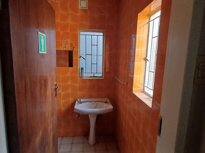 A functional office restroom featuring a pedestal washbasin, orange ceramic wall tiling, and security bars on the windows.