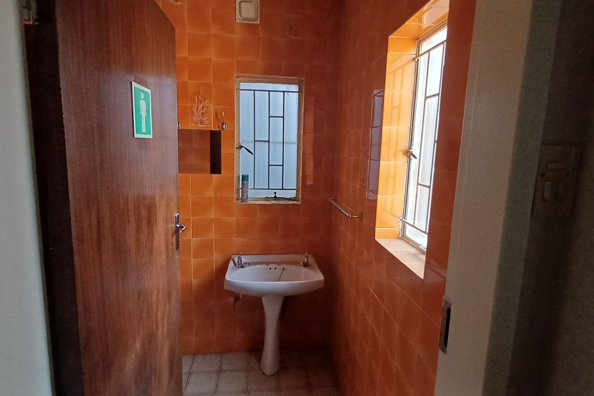 A functional office restroom featuring a pedestal washbasin, orange ceramic wall tiling, and security bars on the windows.