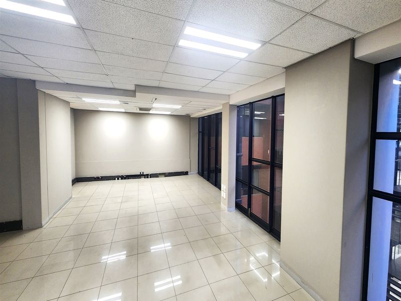 A bright, empty office space featuring tiled flooring, a suspended ceiling with recessed lighting, and large windows with black metal frames.