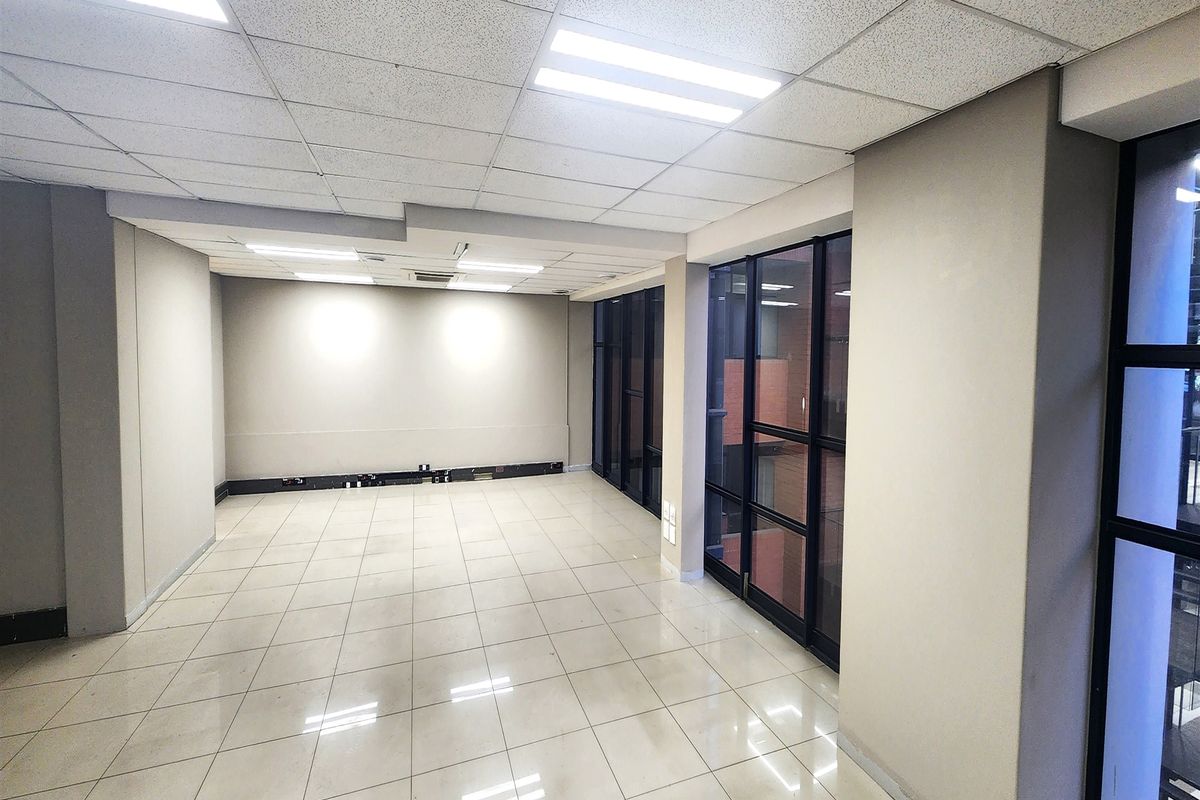 A bright, empty office space featuring tiled flooring, a suspended ceiling with recessed lighting, and large windows with black metal frames.