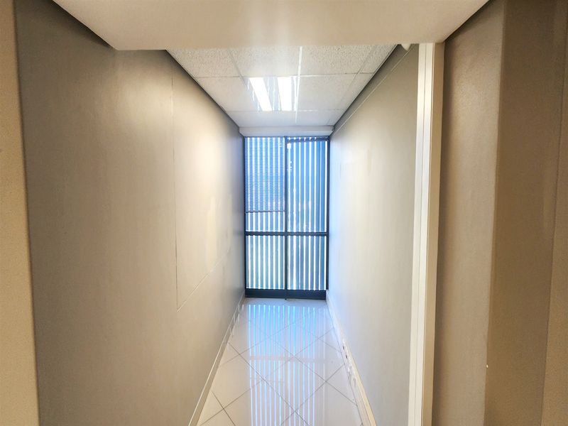A clean, empty office hallway featuring tiled flooring, a suspended ceiling with recessed lighting, and vertical blinds on the window.