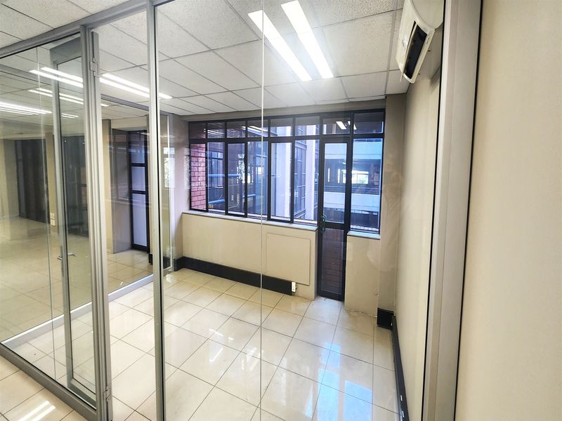 A bright, empty office space featuring glass partition walls, tiled flooring, a suspended ceiling, and a wall-mounted air conditioning unit.