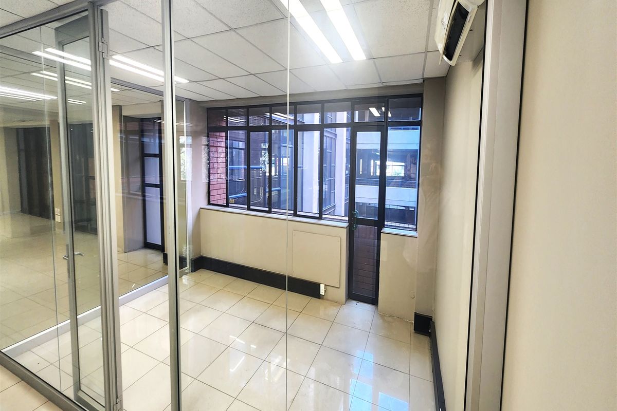 A bright, empty office space featuring glass partition walls, tiled flooring, a suspended ceiling, and a wall-mounted air conditioning unit.
