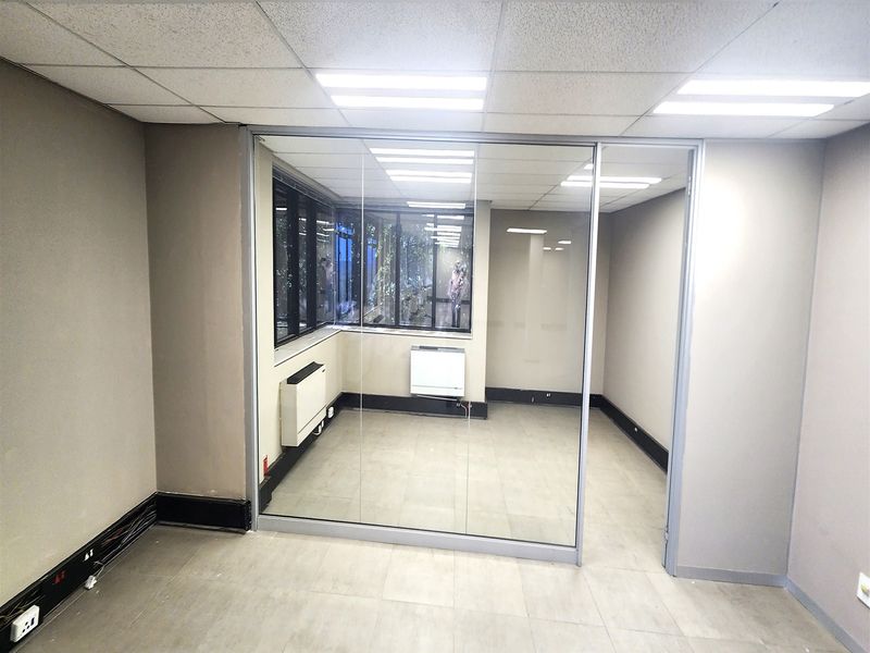 A vacant office space featuring a glass partition wall, tiled flooring, suspended ceiling with recessed lighting, and wall-mounted HVAC units.