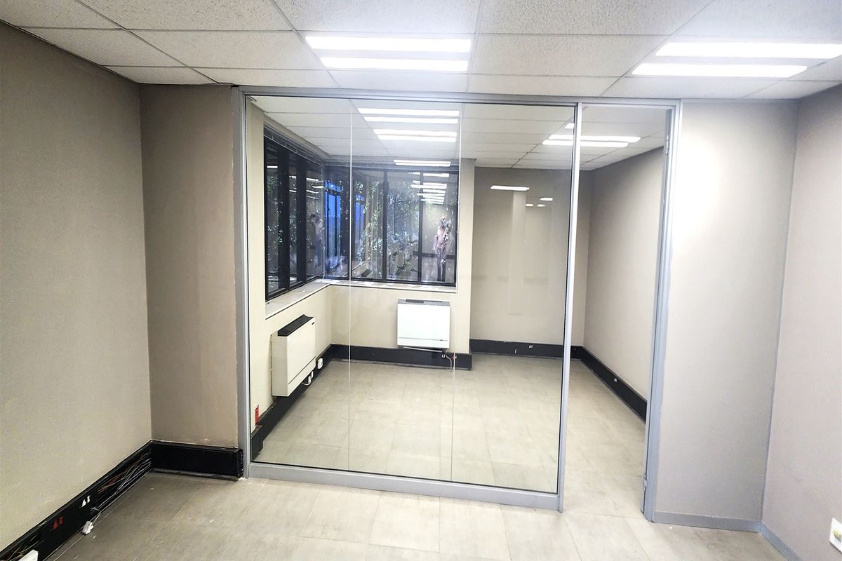 A vacant office space featuring a glass partition wall, tiled flooring, suspended ceiling with recessed lighting, and wall-mounted HVAC units.