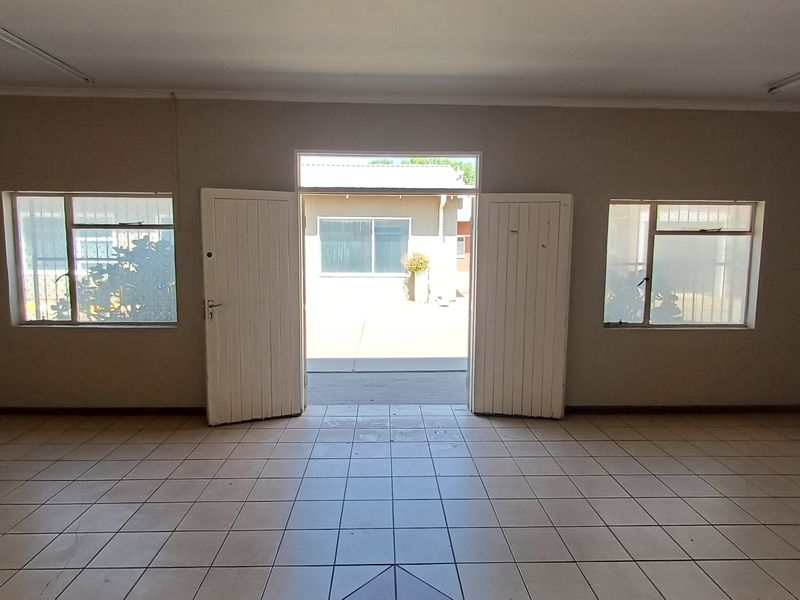 An empty, open-plan office space featuring tiled flooring, fluorescent lighting, and double doors leading to an exterior courtyard area.