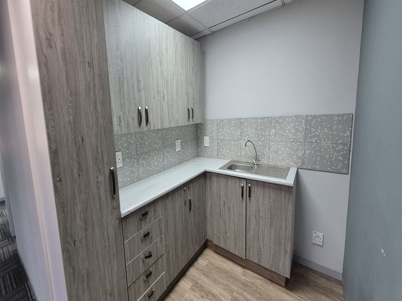 A compact office kitchenette featuring modern wood-look laminate cabinetry, a stainless steel sink, and a decorative tiled backsplash.