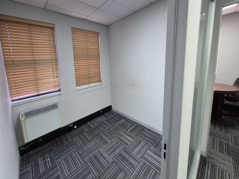 A compact, empty office space featuring carpet tiles, a suspended ceiling, wall-mounted heater, and venetian blinds, viewed through a glass partition door.
