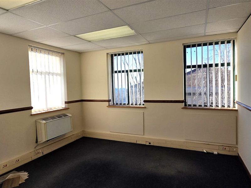 A vacant office space featuring a suspended ceiling, recessed lighting, wall-mounted HVAC unit, and large windows with vertical blinds.