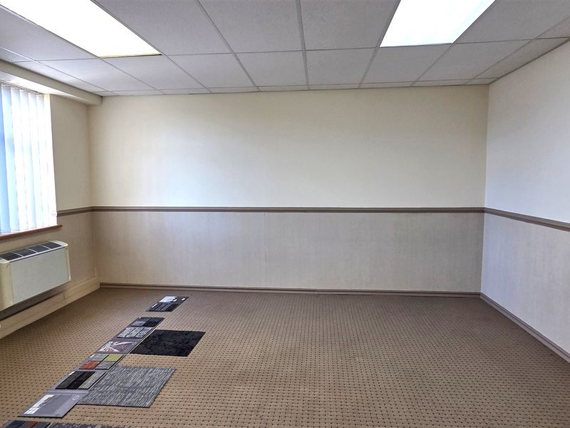 A vacant office space featuring a drop ceiling, recessed lighting, wall-mounted HVAC unit, and neutral carpet flooring with chair rail detailing.