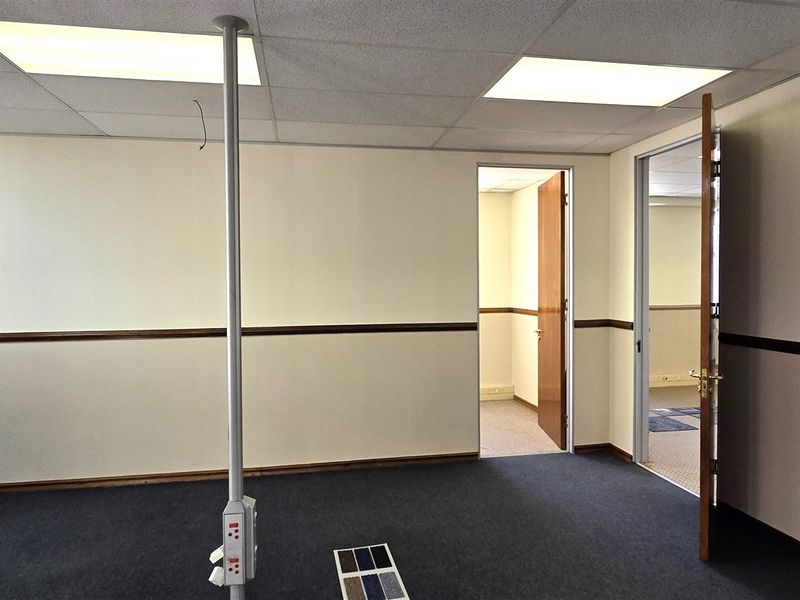 A vacant office space featuring a suspended ceiling, recessed lighting, carpeted flooring, and a central power pole with visible wiring.