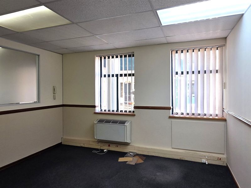 An empty office space featuring a suspended ceiling, recessed lighting, wall-mounted air conditioning, and vertical blinds.