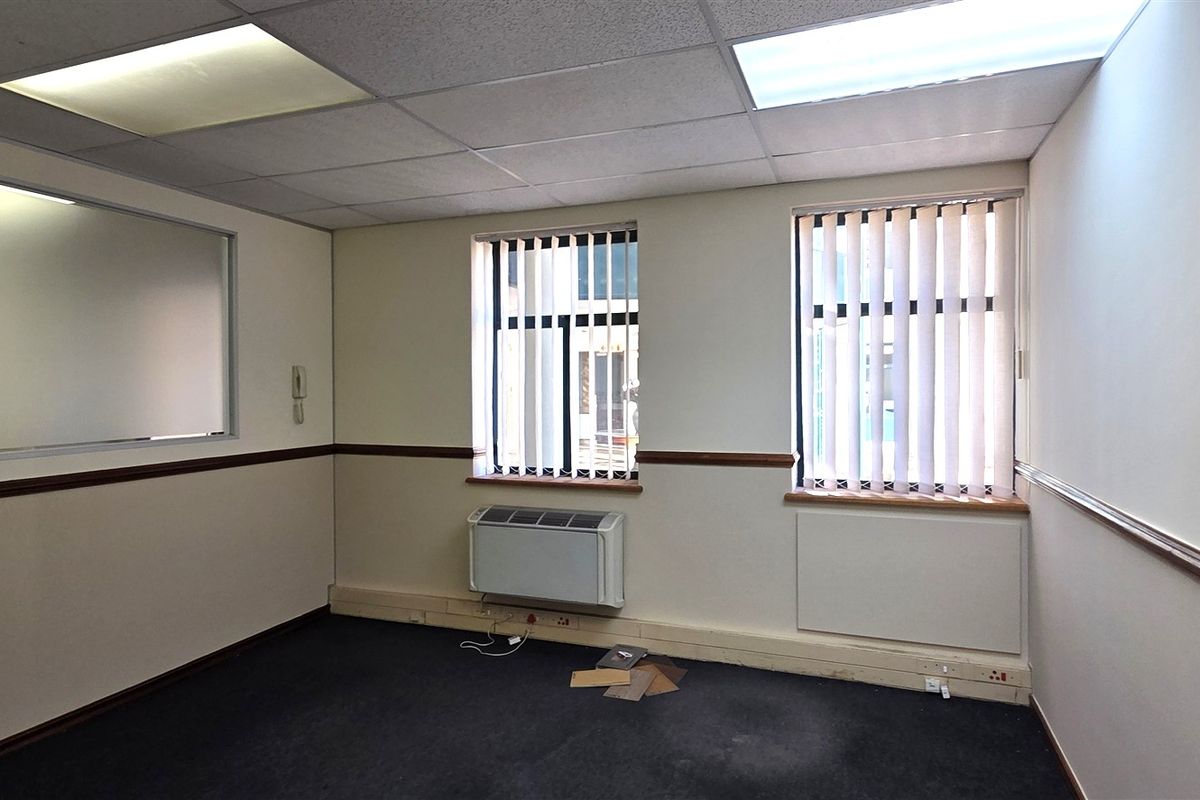 An empty office space featuring a suspended ceiling, recessed lighting, wall-mounted air conditioning, and vertical blinds.