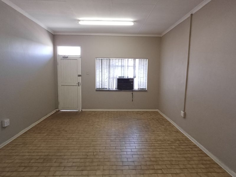 A vacant office space featuring tiled flooring, a window-mounted air conditioning unit, and neutral wall finishes, suitable for a professional workspace.