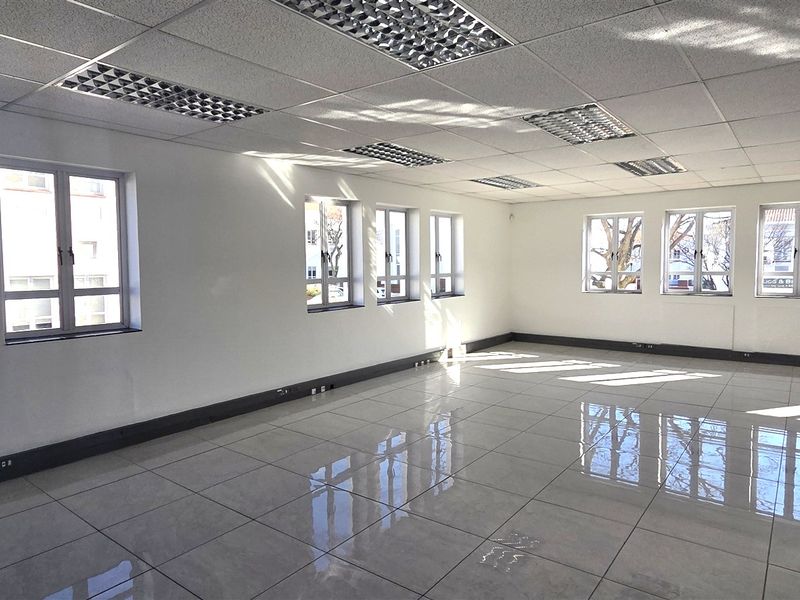 A bright, open-plan office space featuring polished tiled flooring, a suspended ceiling with recessed lighting, and multiple windows providing ample natural light.