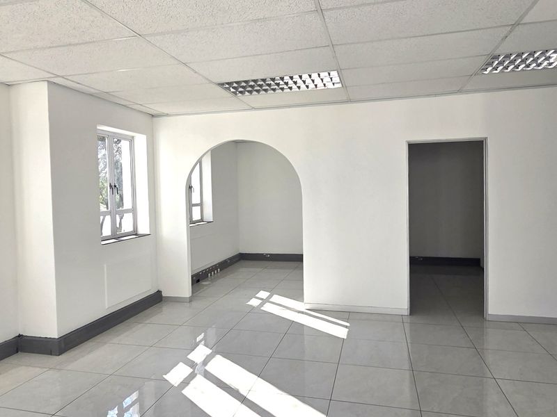 A bright, vacant office space featuring tiled flooring, a suspended ceiling with recessed lighting, and an architectural archway connecting to an adjacent room.