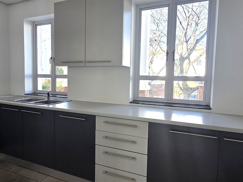 A clean, modern office kitchen area featuring two-tone cabinetry, light countertops, stainless steel sink, and ample natural light from dual windows.