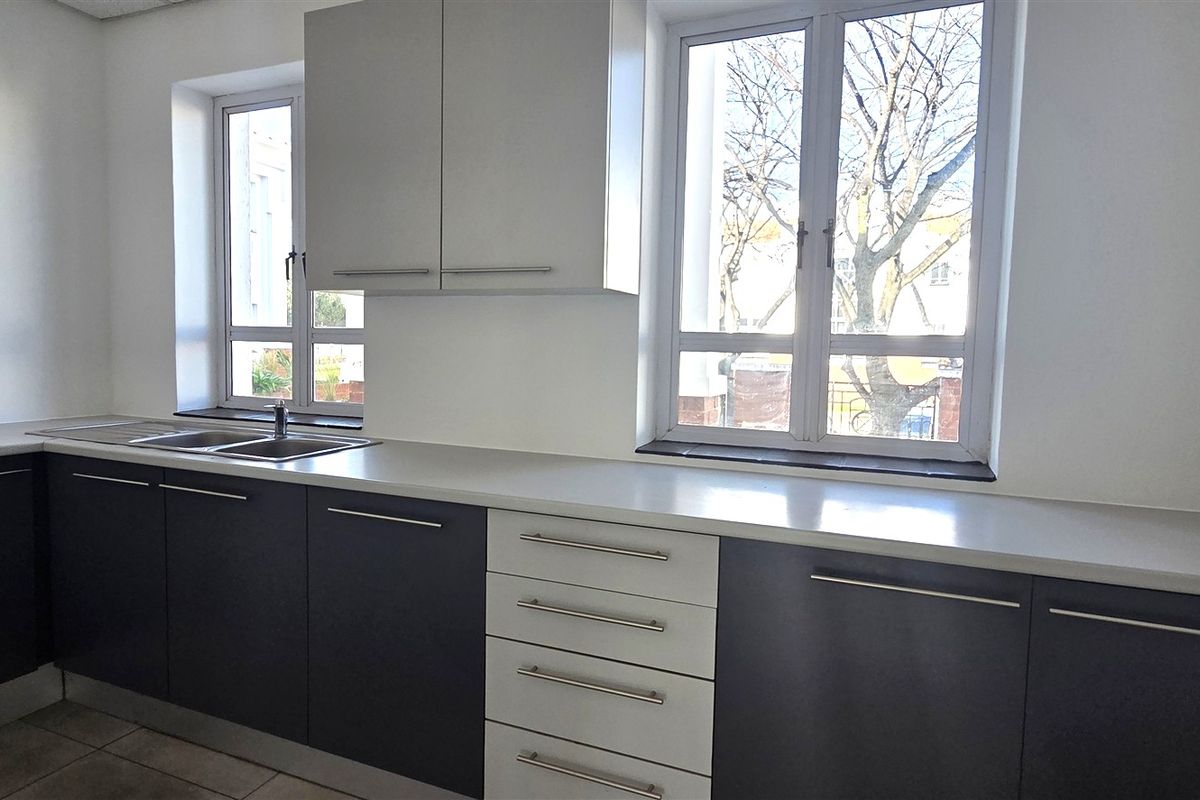 A clean, modern office kitchen area featuring two-tone cabinetry, light countertops, stainless steel sink, and ample natural light from dual windows.