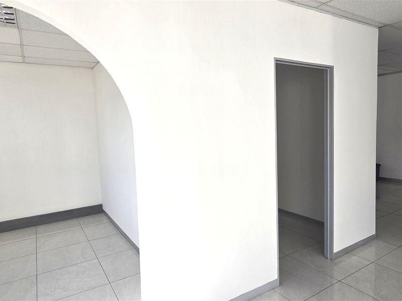 A bright, vacant office space featuring neutral tiled flooring, a suspended ceiling, and an architectural archway connecting two distinct work areas.