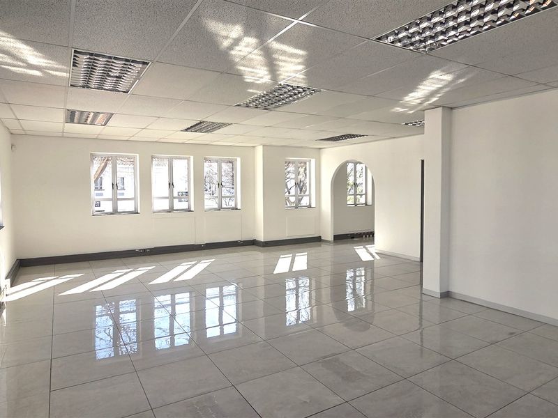 A bright, open-plan office space featuring polished tiled flooring, a suspended ceiling with recessed lighting, and multiple windows providing ample natural light.