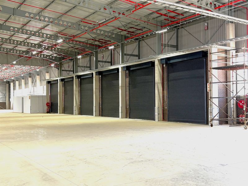 Spacious industrial warehouse interior featuring multiple roller shutter doors, exposed steel roof trusses, and a fire sprinkler system with polished concrete flooring.