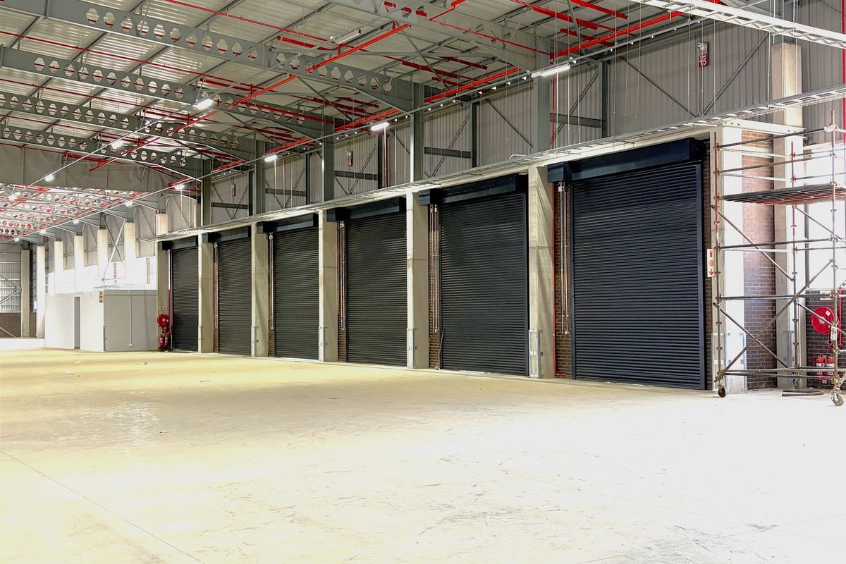 Spacious industrial warehouse interior featuring multiple roller shutter doors, exposed steel roof trusses, and a fire sprinkler system with polished concrete flooring.