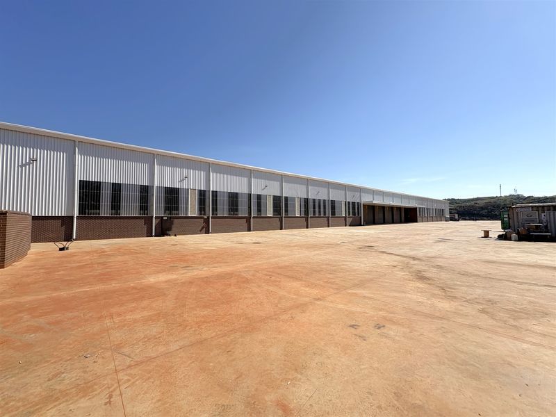 A wide-angle view of a large industrial warehouse exterior featuring corrugated metal cladding, brick base, and an expansive concrete yard area.
