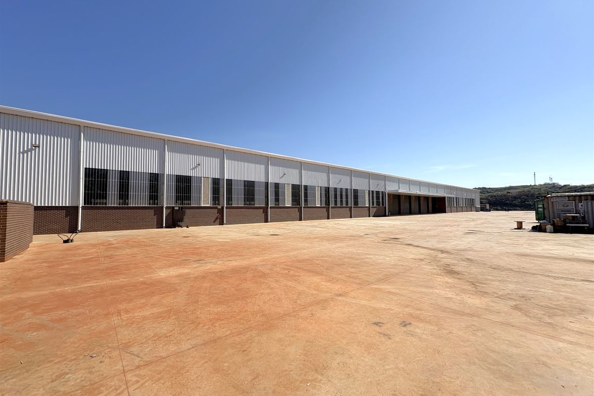 A wide-angle view of a large industrial warehouse exterior featuring corrugated metal cladding, brick base, and an expansive concrete yard area.