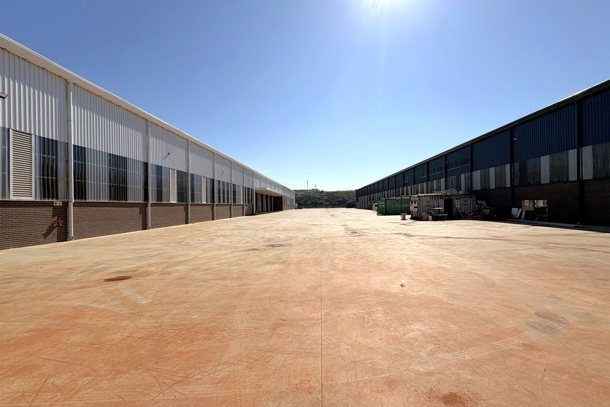 Expansive industrial concrete yard situated between two large warehouse structures with metal cladding and brick facade finishes.