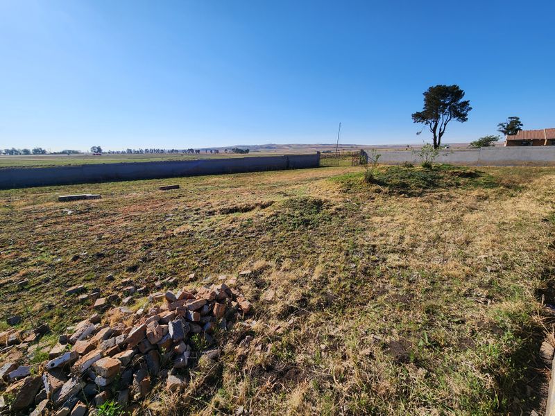 A view of the vacant commercial land featuring a perimeter wall, metal gate, and open grass area with some scattered debris.