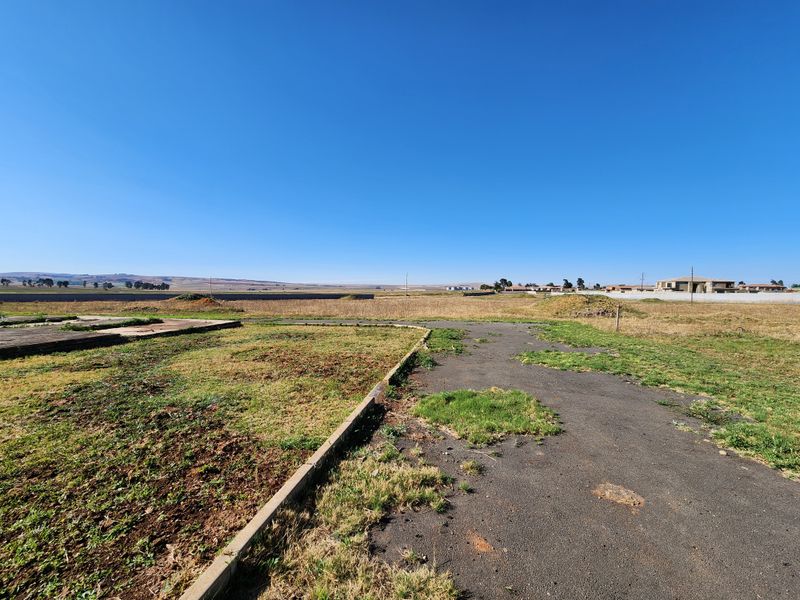 A wide-angle view of a vacant commercial land plot featuring existing asphalt paving and concrete curbing under a clear blue sky.
