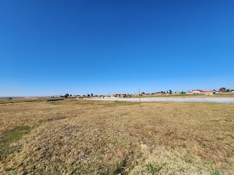 A wide-angle view of a vacant commercial land plot featuring dry grass and a perimeter boundary wall in the distance.